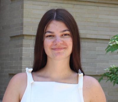 Person with long brown straight hair and a white top smiles at the camera straight on