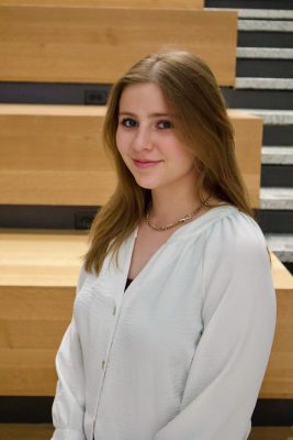 Person with long light brown hair and and white top smiles at the camera while standing sideways