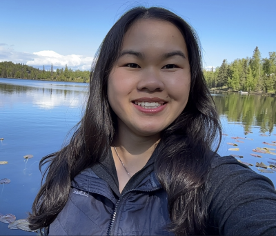 smiling person with long dark brown hair stands in front of a lake or lagoon